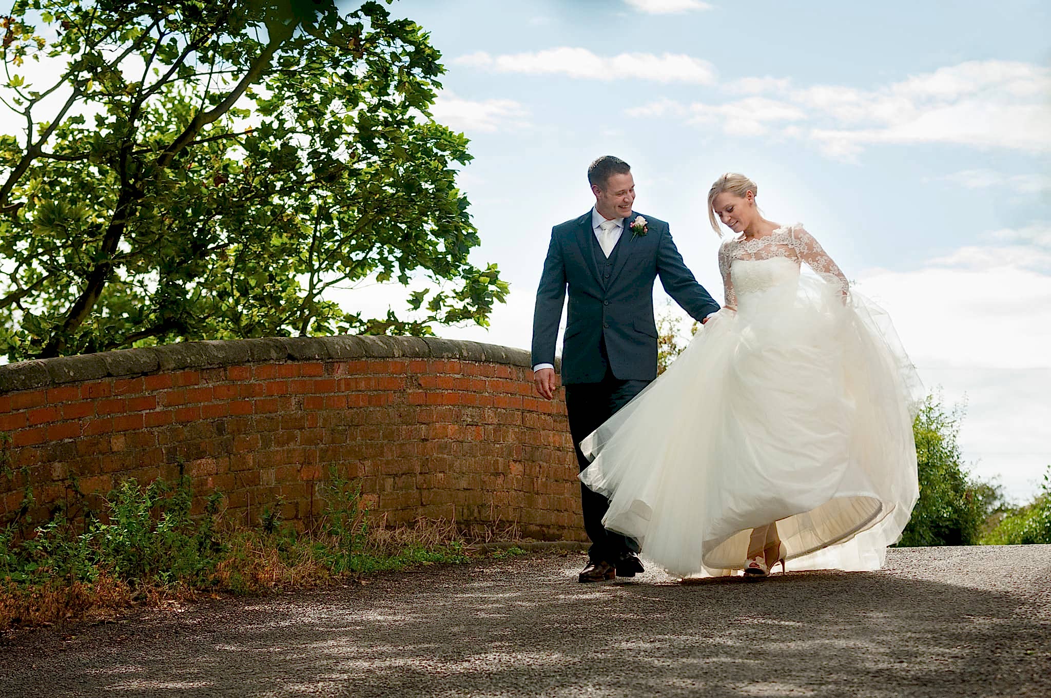 Groom helping the bride with her dress as they walk over a bridge with red brick walls outside Dodmoor House