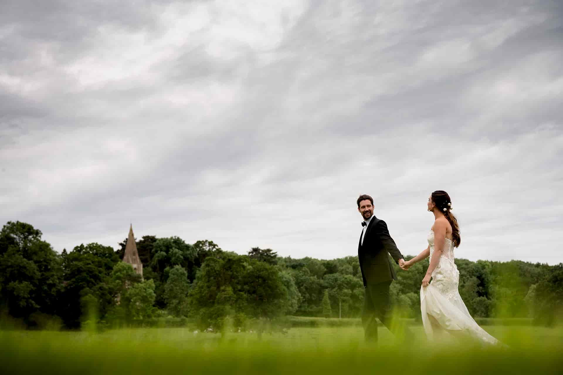 Bride and groom walking through field with church in shot