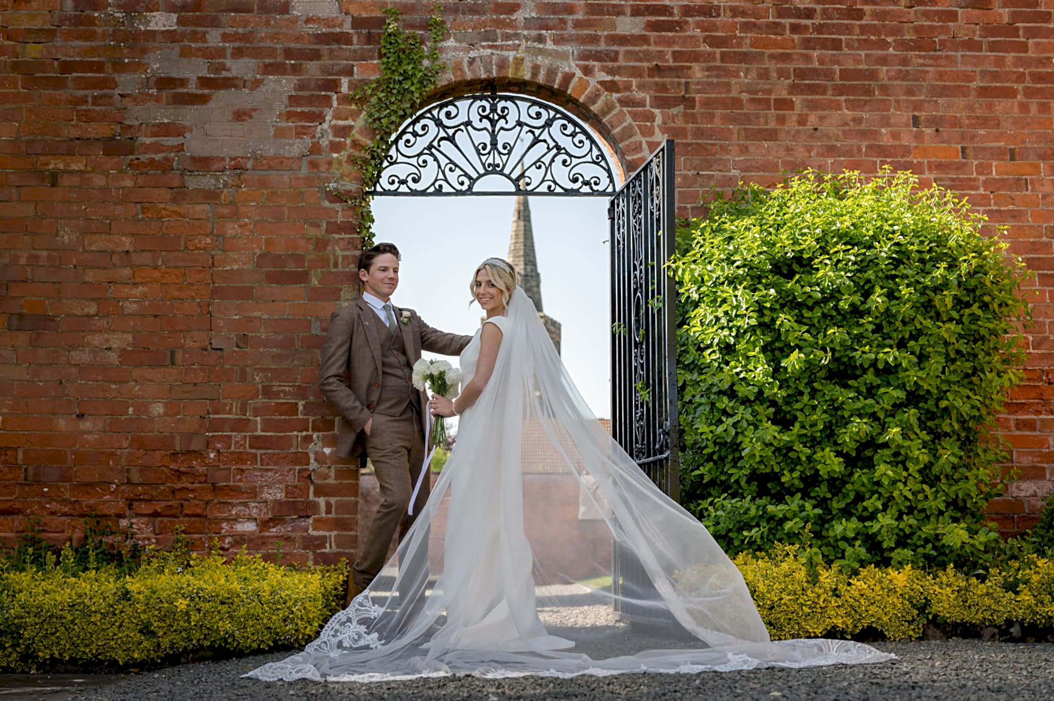 Bride and groom in front of a wrought iron gate with the spire of Sutton Bonington church in the background