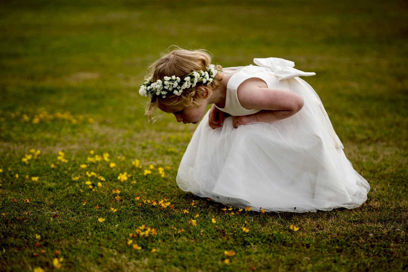 A flower girl in a white dress and flower crown bending down to look closely at buttercups in the lawn