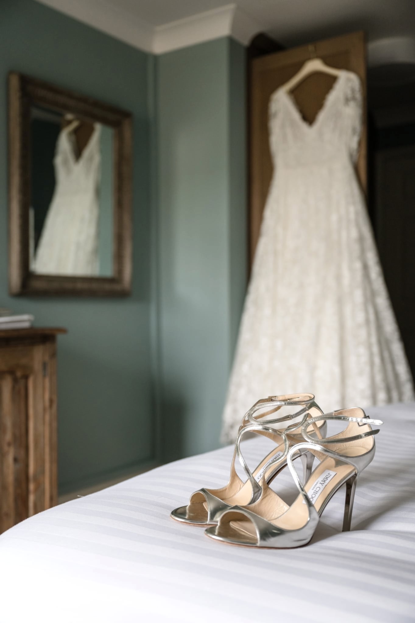 Bride's shoes on the end of the bed with her dress hanging in the background