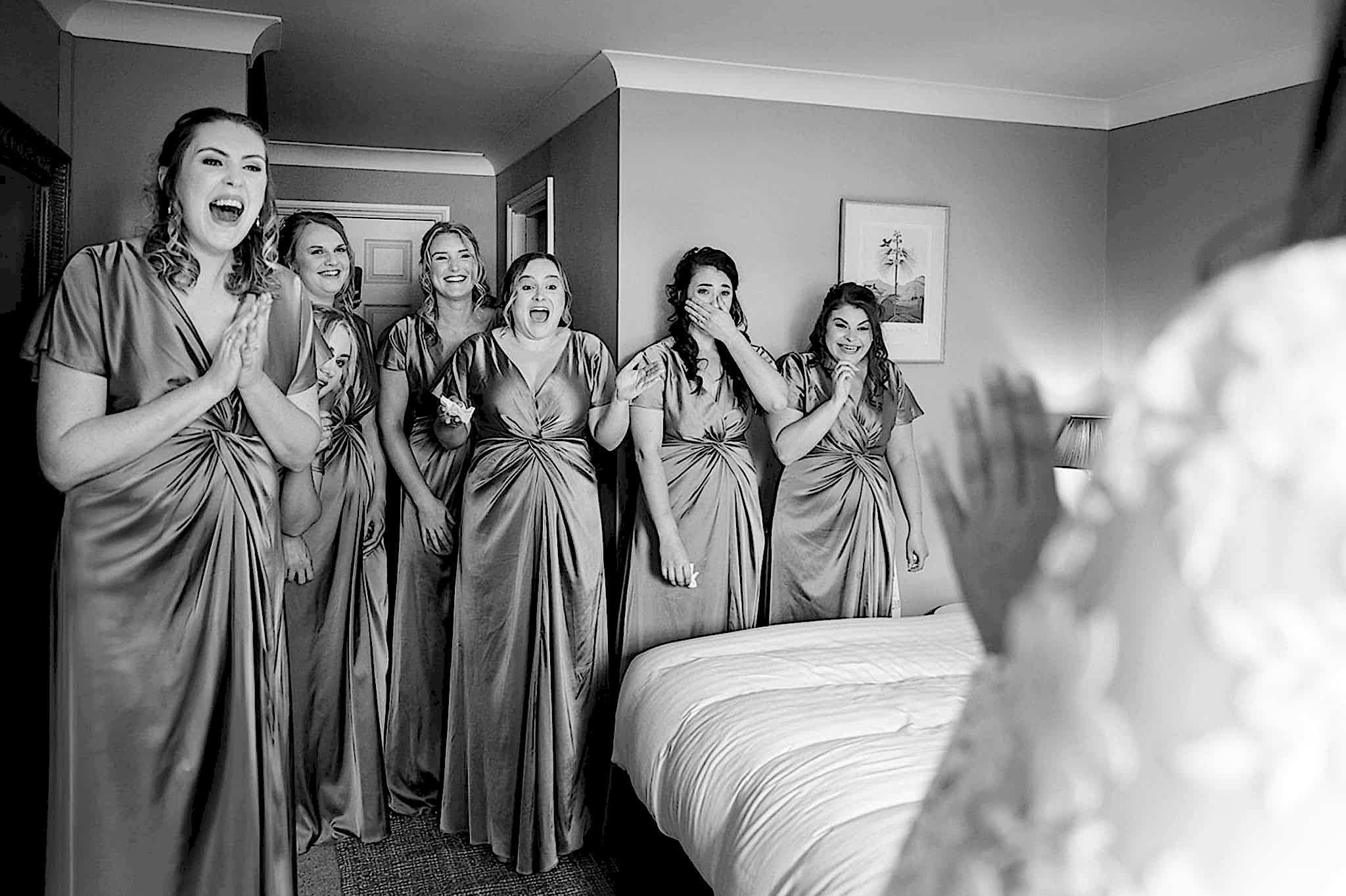 7 bridesmaids with various reactions on their faces as they see the bride in her dress for the first time