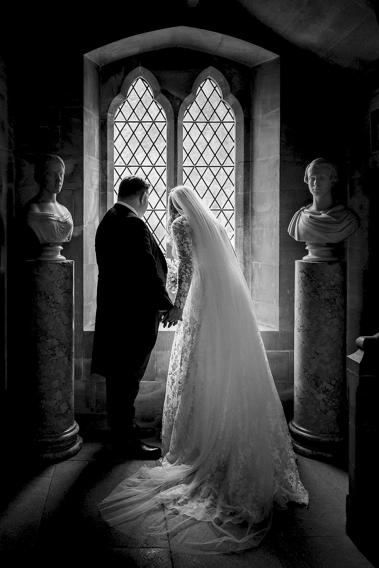 Bride and groom looking out of a beautiful window flanked by marble busts at Exton Park chapel