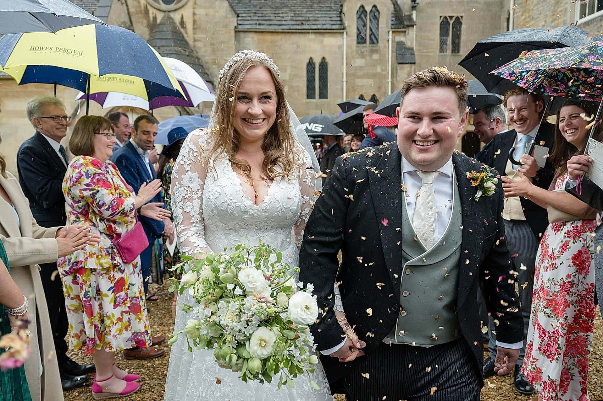 Bride and groom laughing as their guests throw confetti in the rain at Exton Park chapel