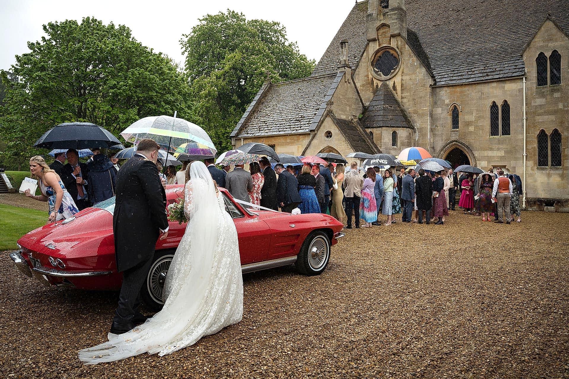 Bride and groom getting into a red vintage wedding car outside Exton Park chapel as all the guests shelter under umbrellas