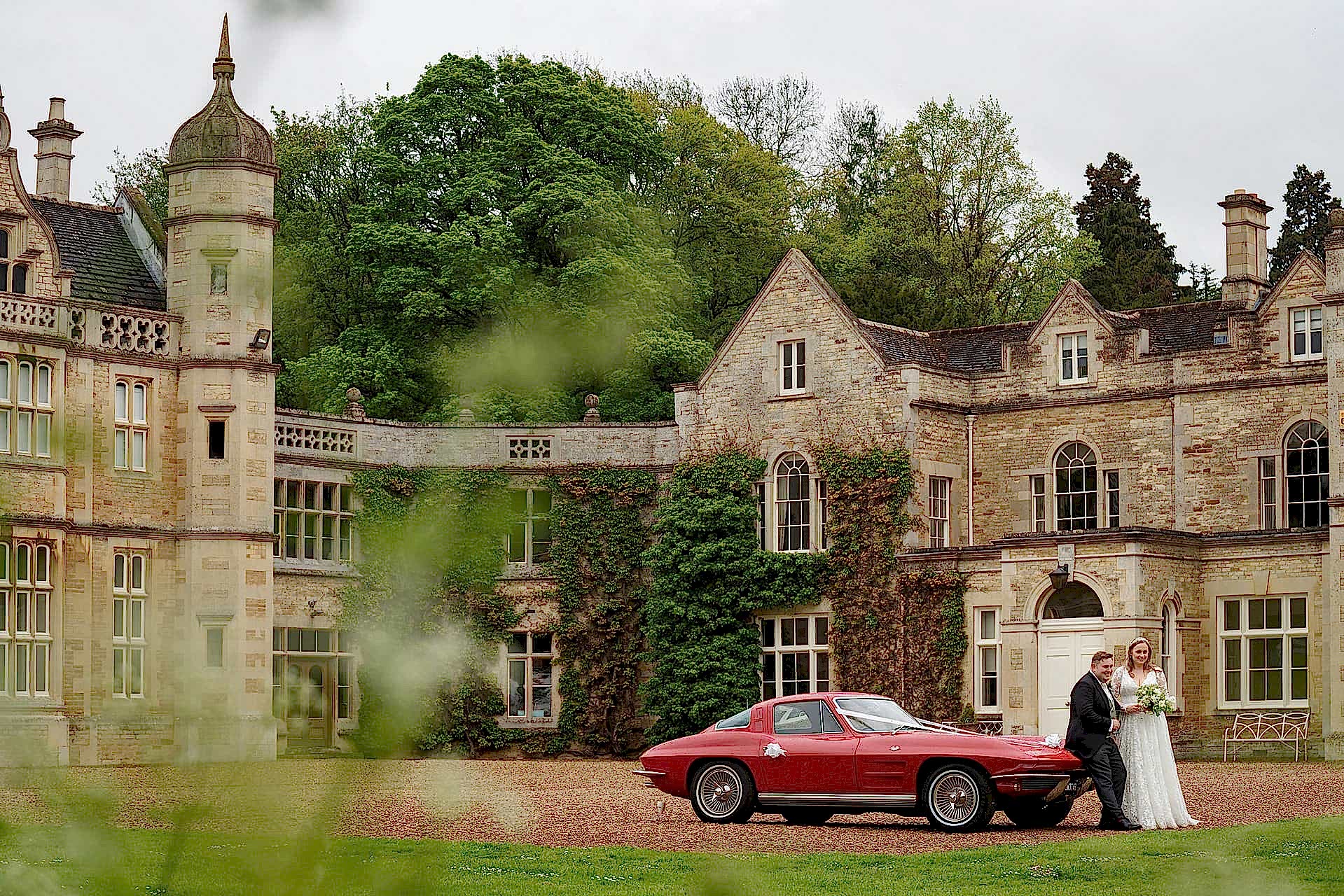 Bride and groom standing by their red vintage wedding car outside Exton Park Hall
