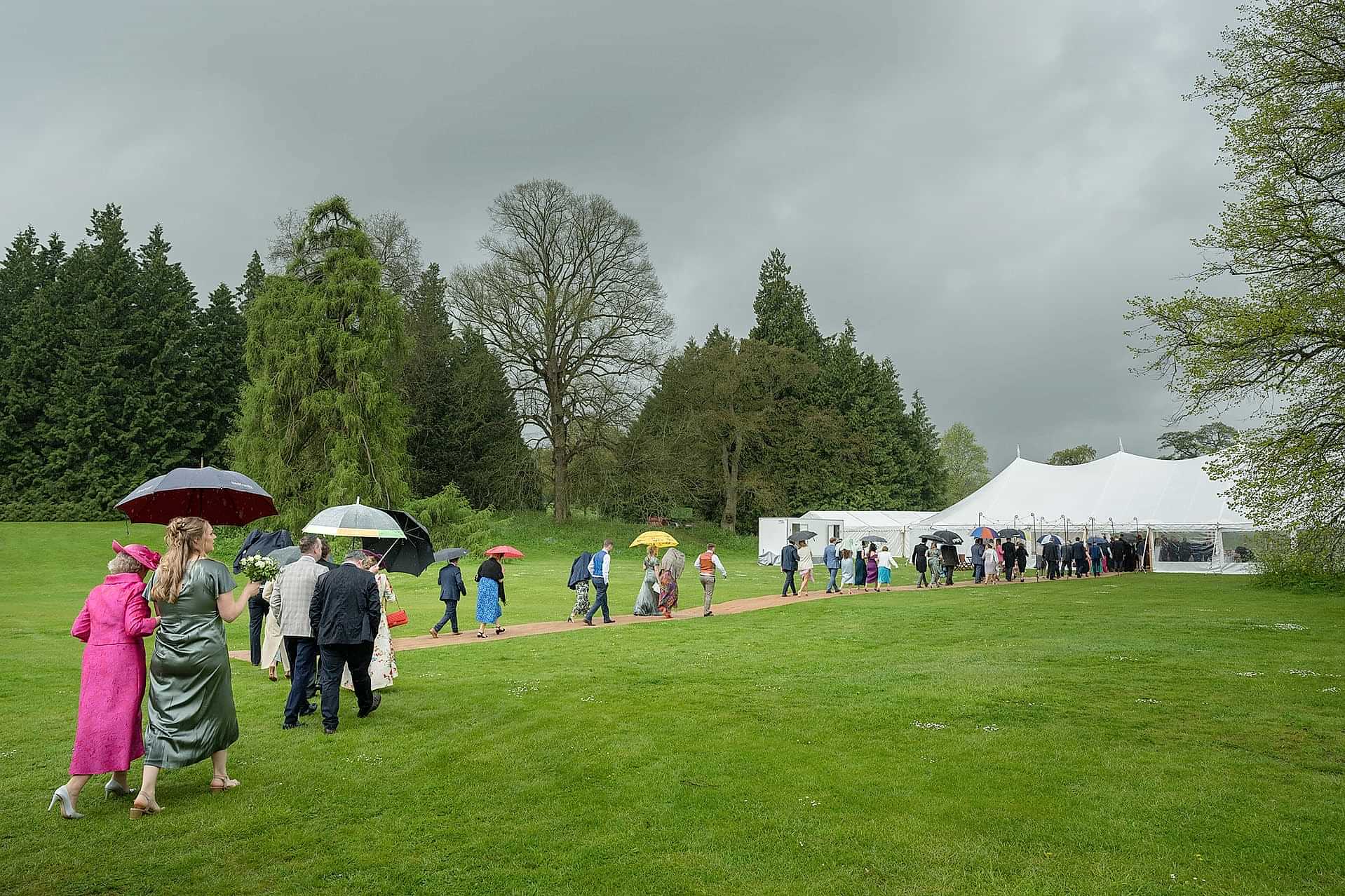 Wedding guests walking to the marquee under umbrellas in the rain at Exton Park