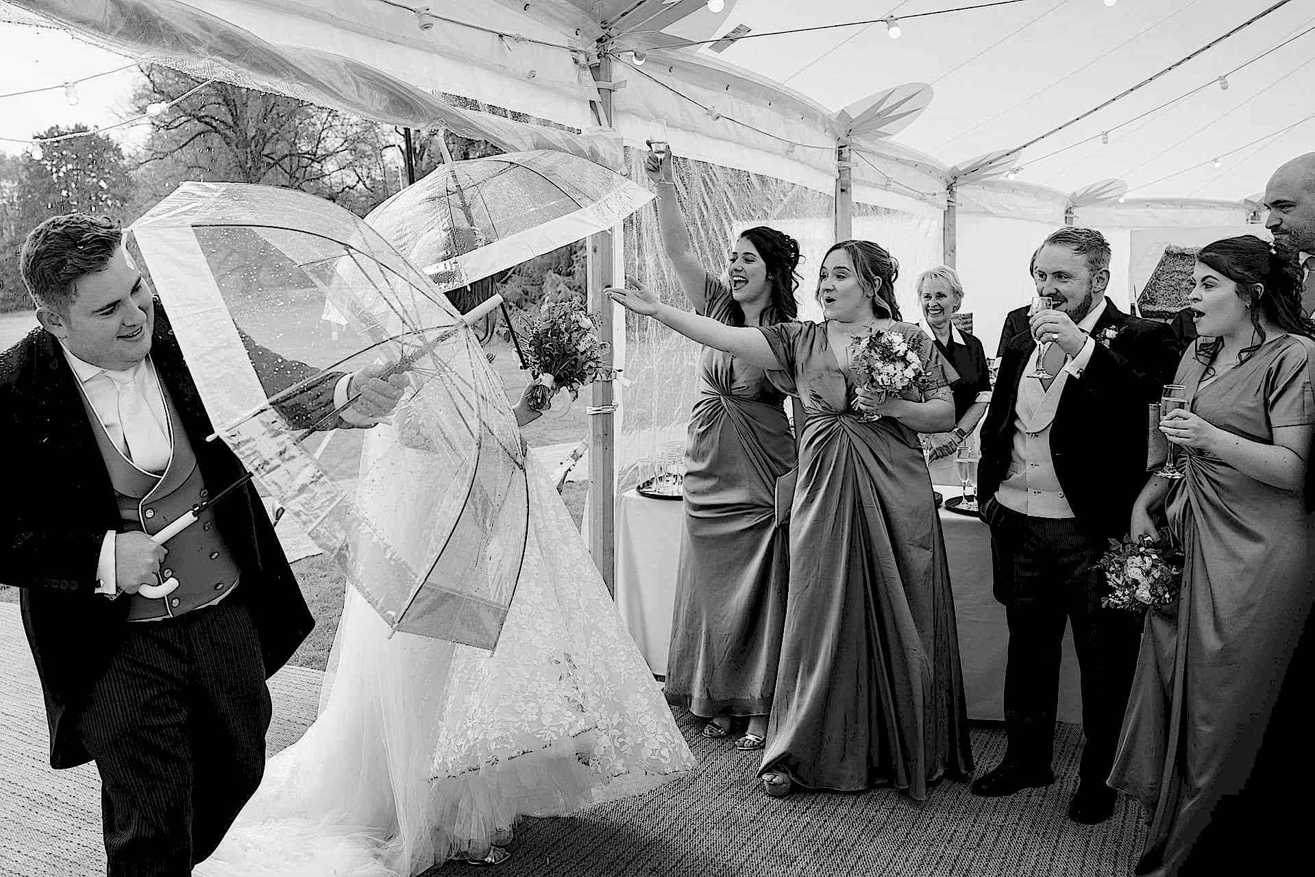 Bridesmaids reaching out to help the bride and groom as they walk into the marquee under umbrellas