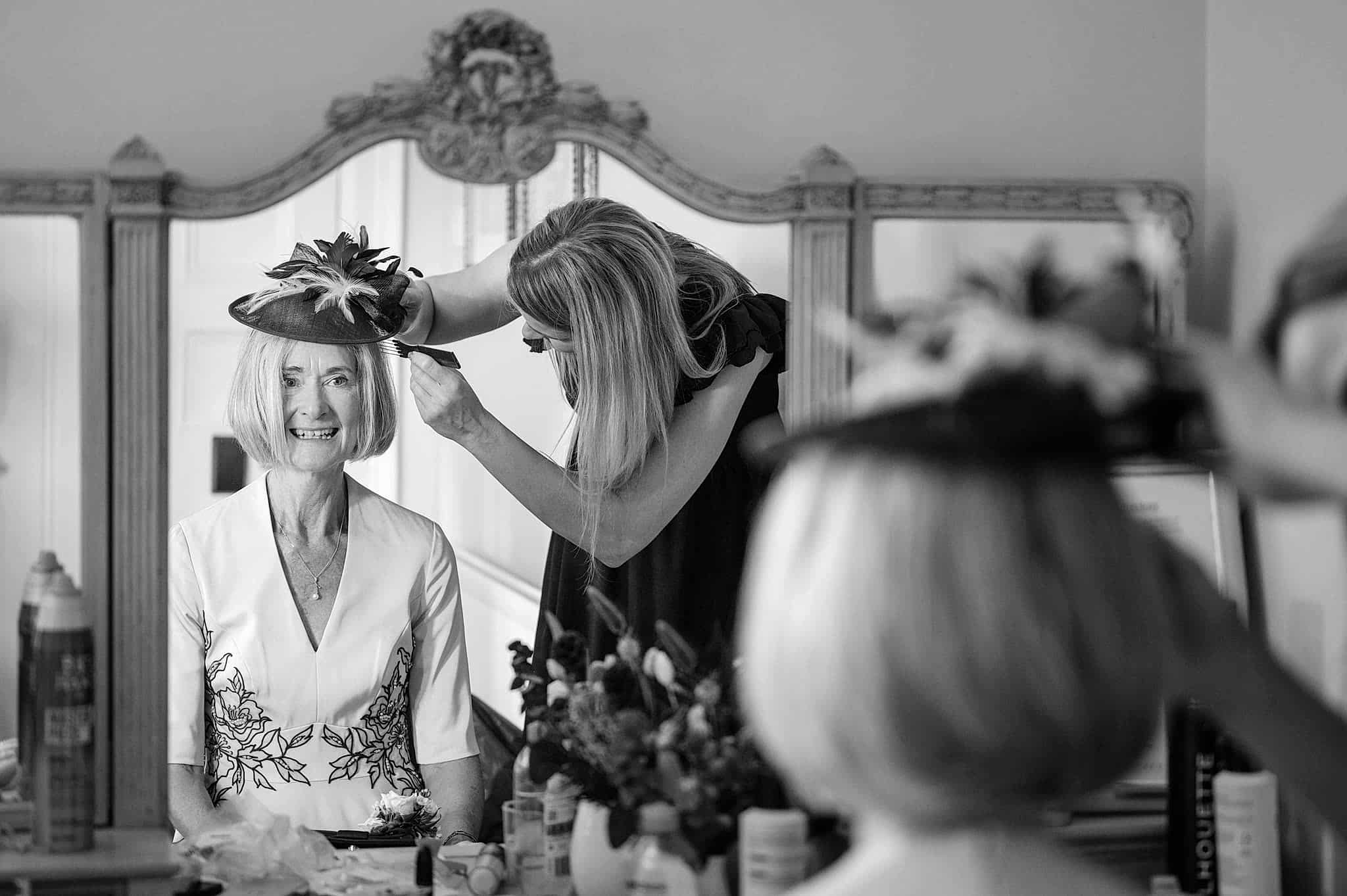 A mirror reflection of a hair stylist adjusting the mother of the groom's hair and hat