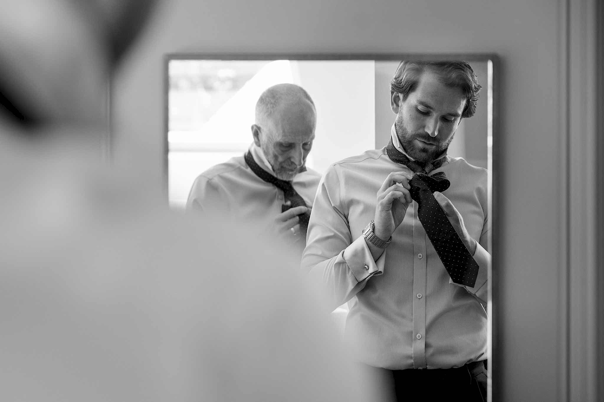 A mirror reflection of a groom and his dad putting on their ties together