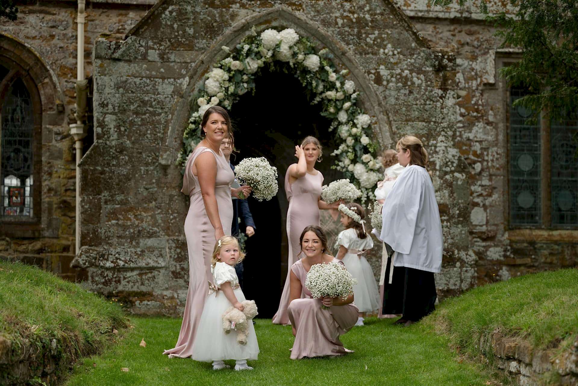 Bridesmaids and flower girls in nude pink dresses waiting outside Fawsley church for the bride to arrive