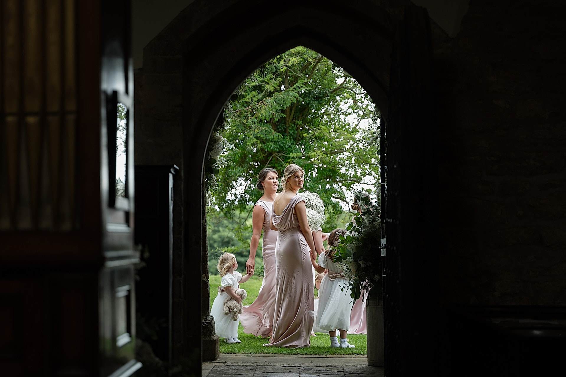 Bridesmaids and flower girls looking through the door into Fawsley church while they wait for the bride to arrive