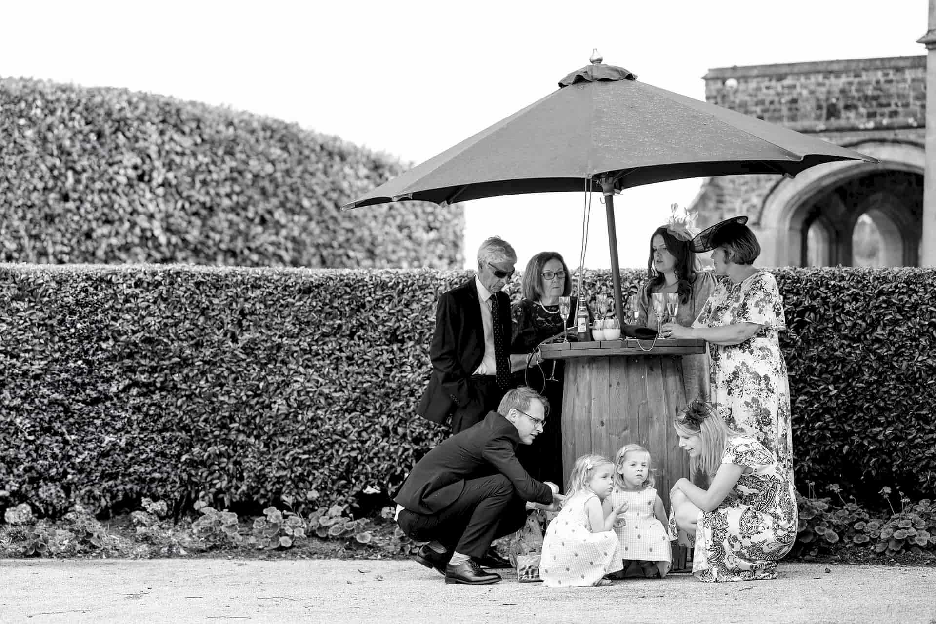 A family gathered around an oak barrel table and sheltering under a parasol as it starts to rain