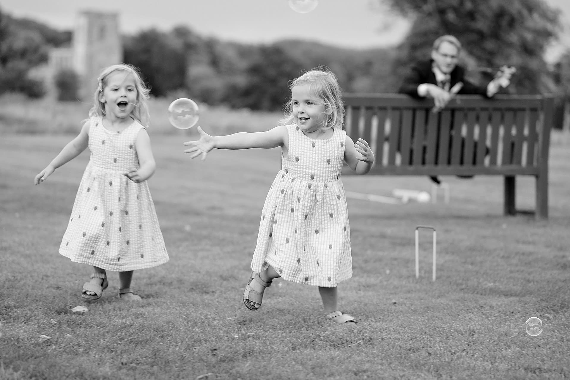 Two young twin girls in matching outfits playing with bubbles with Fawsley church in the background