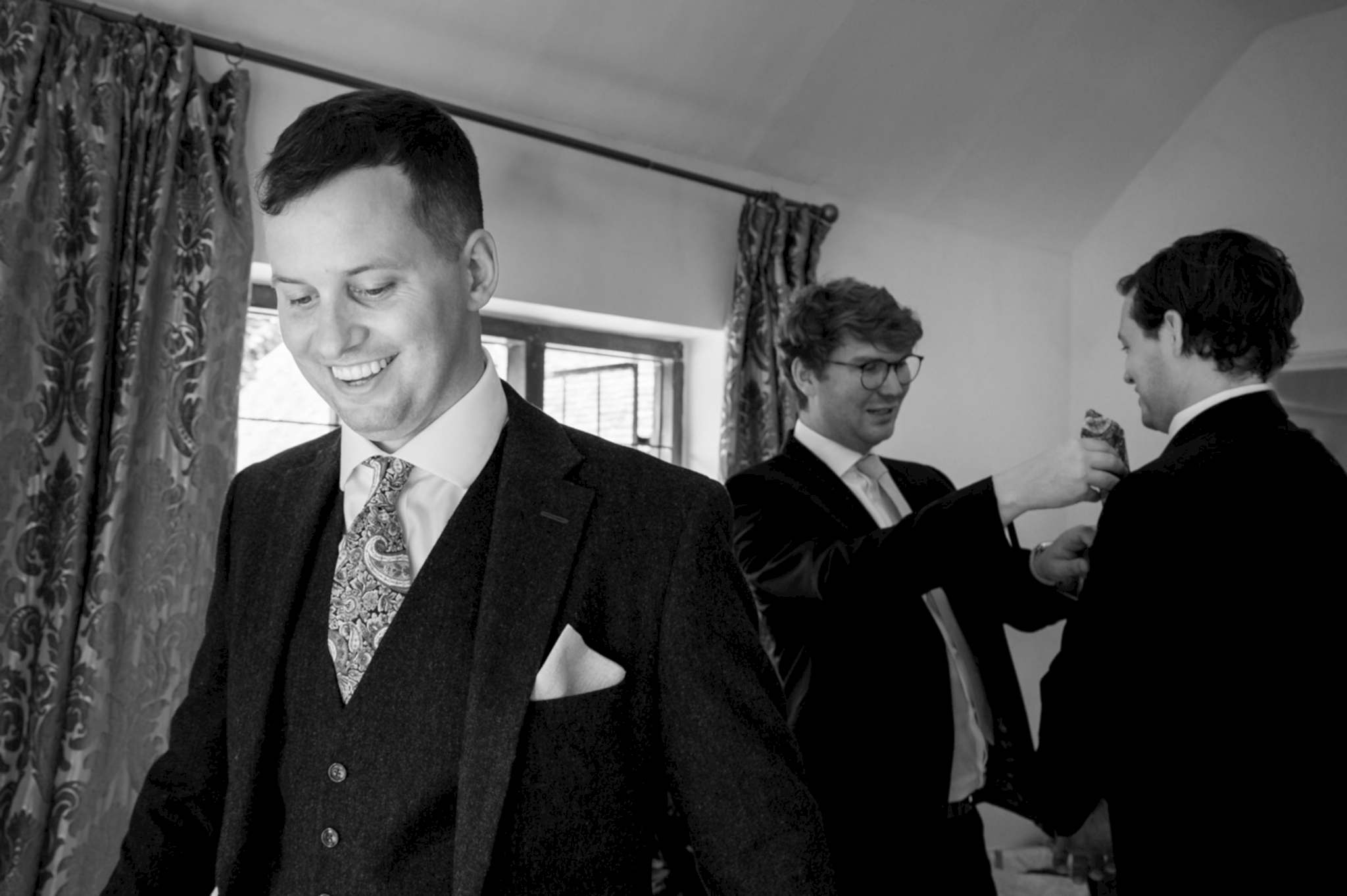 The groom smiling in the foreground as two ushers fold pocket squares behind him