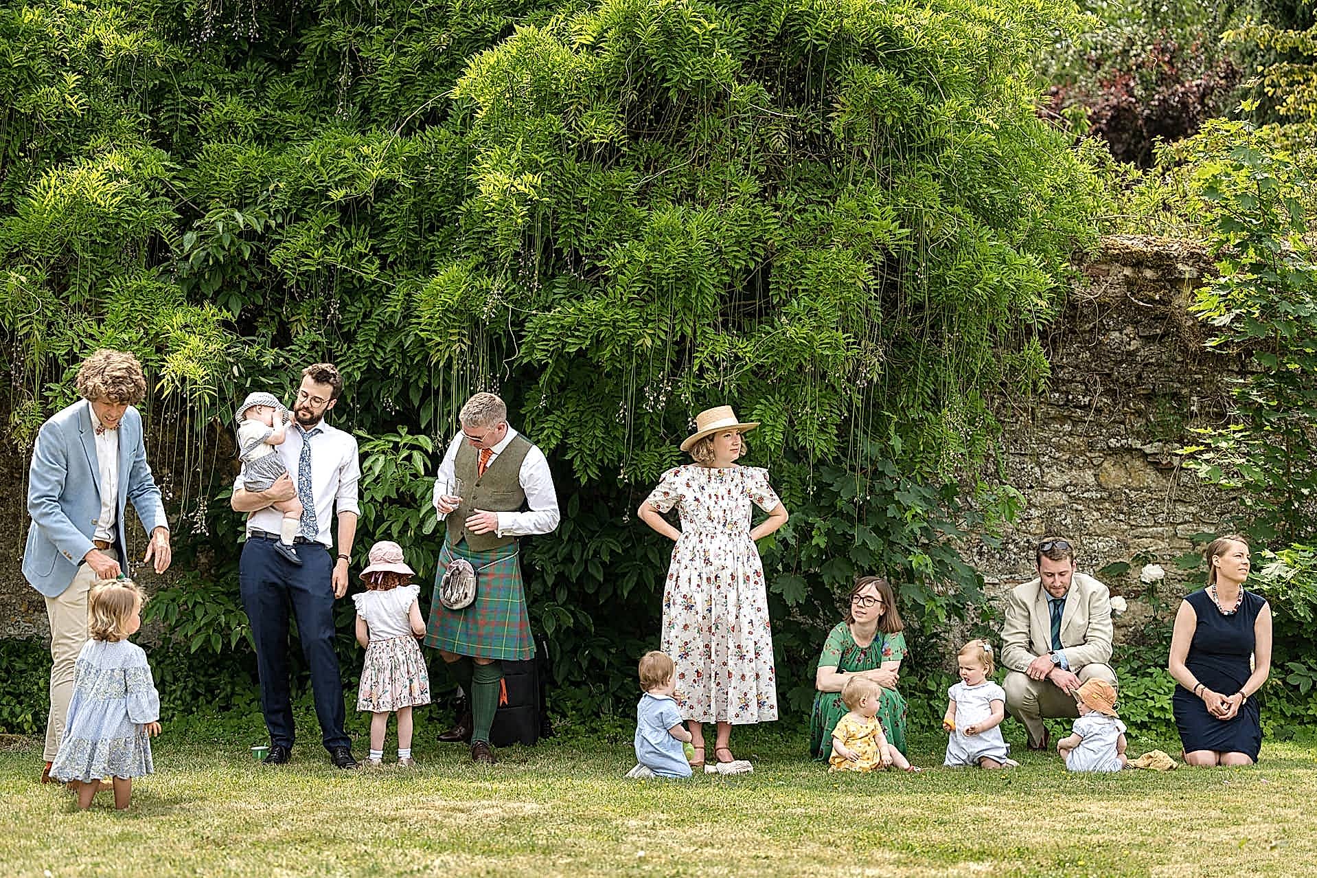 A row of wedding guests taking shelter from the sun in the shade of a wysteria-clad stone wall at Notley Abbey