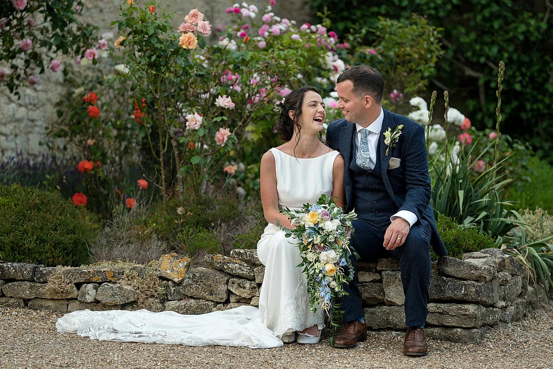 Bride and groom sitting on a low stone wall in front of flowering rose bushes at Notley Abbey