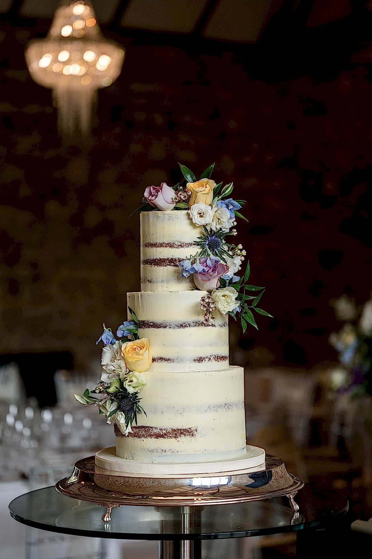 A three-tier semi-naked wedding cake with fresh flowers displayed in the Monk's Refectory at Notley Abbey
