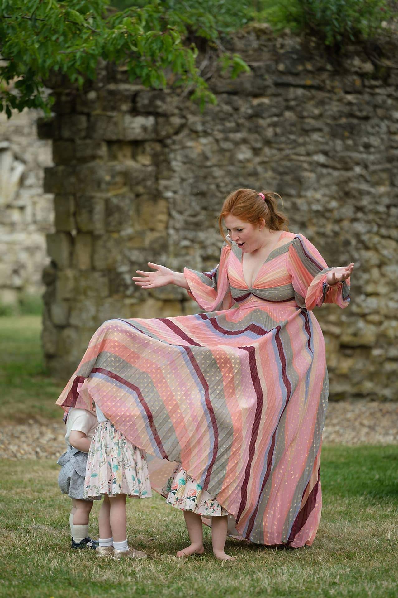 Three small children playing hide and seek under their mum's long floaty dress