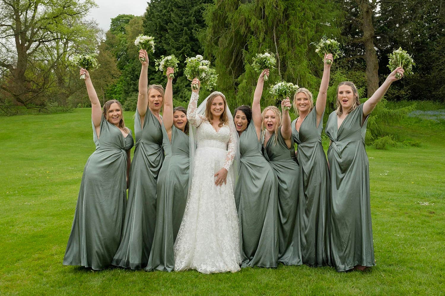 Group photo of bridesmaids in sage green dresses holding their bouquets high in the air